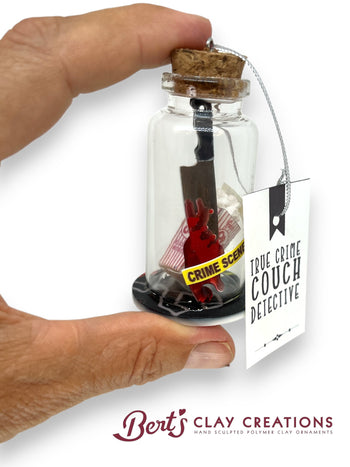 Glass bottle with cork lid containing a small sculpture labeled 'True Crime Couch Detective' by Berts Clay Creations.